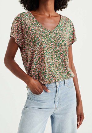 Vrouw draagt een groene, korte mouwen, V-hals top met bloemenmotief, ingestopt in lichtblauwe, hoog taille jeans, met hand in de zak.