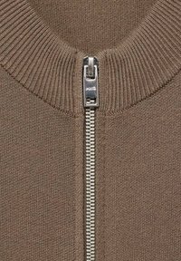 Close-up van een taupe gebreid kledingstuk met een zilveren metalen rits, een geribde kraag en in elkaar grijpende tanden.