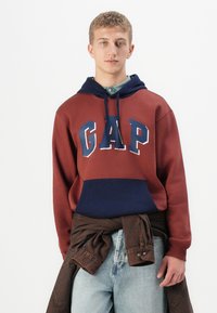 Jeune homme portant un hoodie maroon et navy de GAP, avec une veste en denim nouée autour de la taille et un jean bleu clair, se tenant devant un fond blanc.