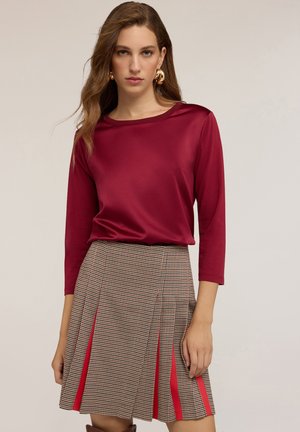 Burgunderfarbener Satin-Top mit dreiviertelärmel, kombiniert mit einem Hahnentritt-Rock mit roten Seitenstreifen und Falten.