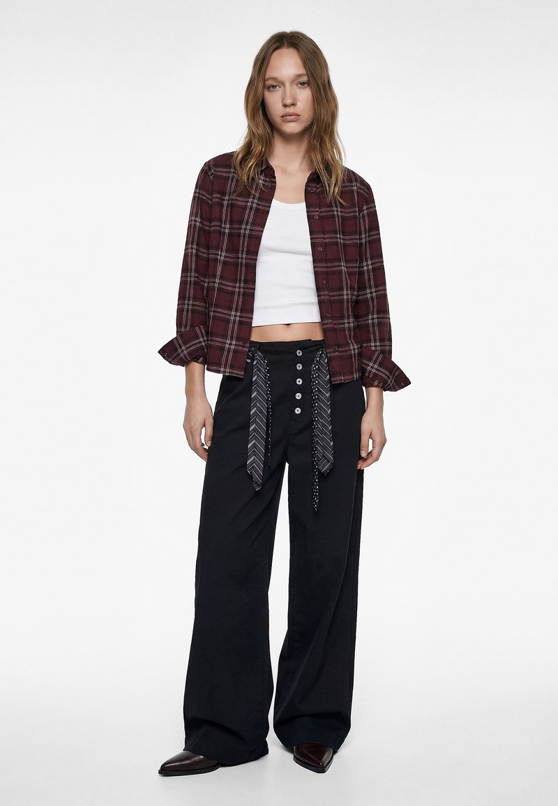 Nuori nainen, jolla on päällään burgundinpunainen ruutupaita valkoisen lyhyen yläosan päällä, mustat leveälahkeiset housut, joissa on napit ja kuvioidut nauhat.