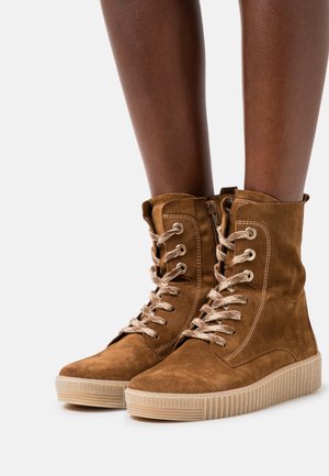 Bottes montantes en daim marron à lacets avec fermeture éclair latérale, portées sur jambes nues, dotées de semelles en caoutchouc à prise texturée.