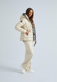 Helle beigefarbene Pufferjacke mit Kapuze, ausgestattet mit einem auffälligen schwarzen Logo-Gurt. Kombiniert mit beigen Hosen und weißen Sneakers. Glatte Textur.