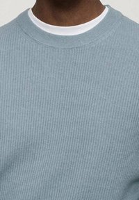 Gros plan sur une personne portant un pull côtelé bleu clair par-dessus un t-shirt blanc à col rond, montrant le cou et la partie supérieure de la poitrine.