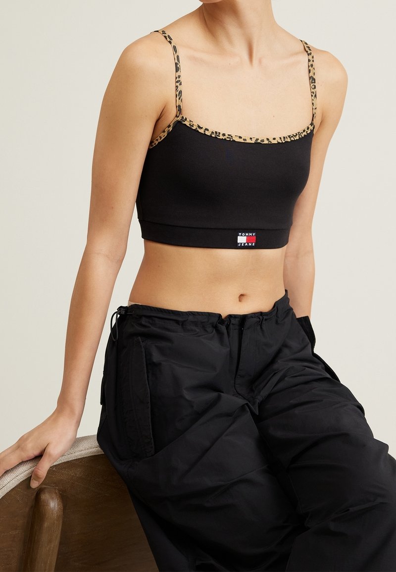 Personne portant un crop top noir avec des bretelles et des bordures à imprimé léopard, et un pantalon noir, assise sur une chaise en bois devant un fond uni.