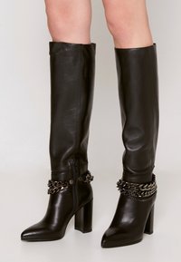 Mustat nahkaiset polvipituiset saappaat, joissa on teräväkärkinen malli, tukeva korko ja ketjulaikku nilkan ympärillä. Varustettu sivuvetoketjulla.