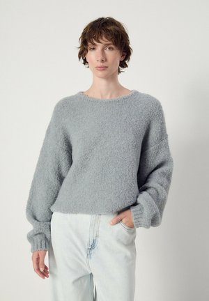 Person mit kurzen lockigen Haaren trägt einen hellgrauen, flauschigen Pullover und hellblaue Jeans und steht mit einer Hand in der Tasche vor einem einfarbigen Hintergrund.