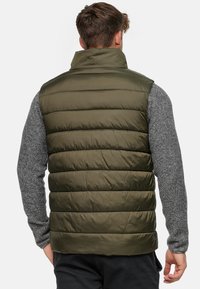 Oliven grøn puffer-vest med høj krave, horisontalt quiltet design og en glat, skinnende tekstur. Båret over en grå, tekstureret langærmet skjorte.