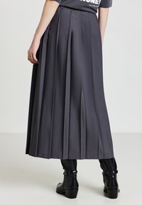 Donna che indossa una gonna midi plissettata grigia con stivaletti neri, in piedi contro uno sfondo grigio chiaro uniforme.