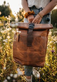 Mochila de cuero marrón con tapa enrollable, correa marrón y un bolsillo delantero con cremallera. Presenta un acabado texturizado y un sutil detalle de logo.