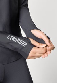 Black athletic long-sleeve top with thumbholes; features "STRONGER" text in gray on the sleeve. Smooth, stretch fabric, fitted design.