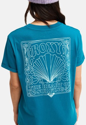 Personne portant un t-shirt bleu canard avec l'inscription « ROXY The Heart of Surfing » et un grand coquillage blanc graphique dans le dos, de dos à l'appareil photo.