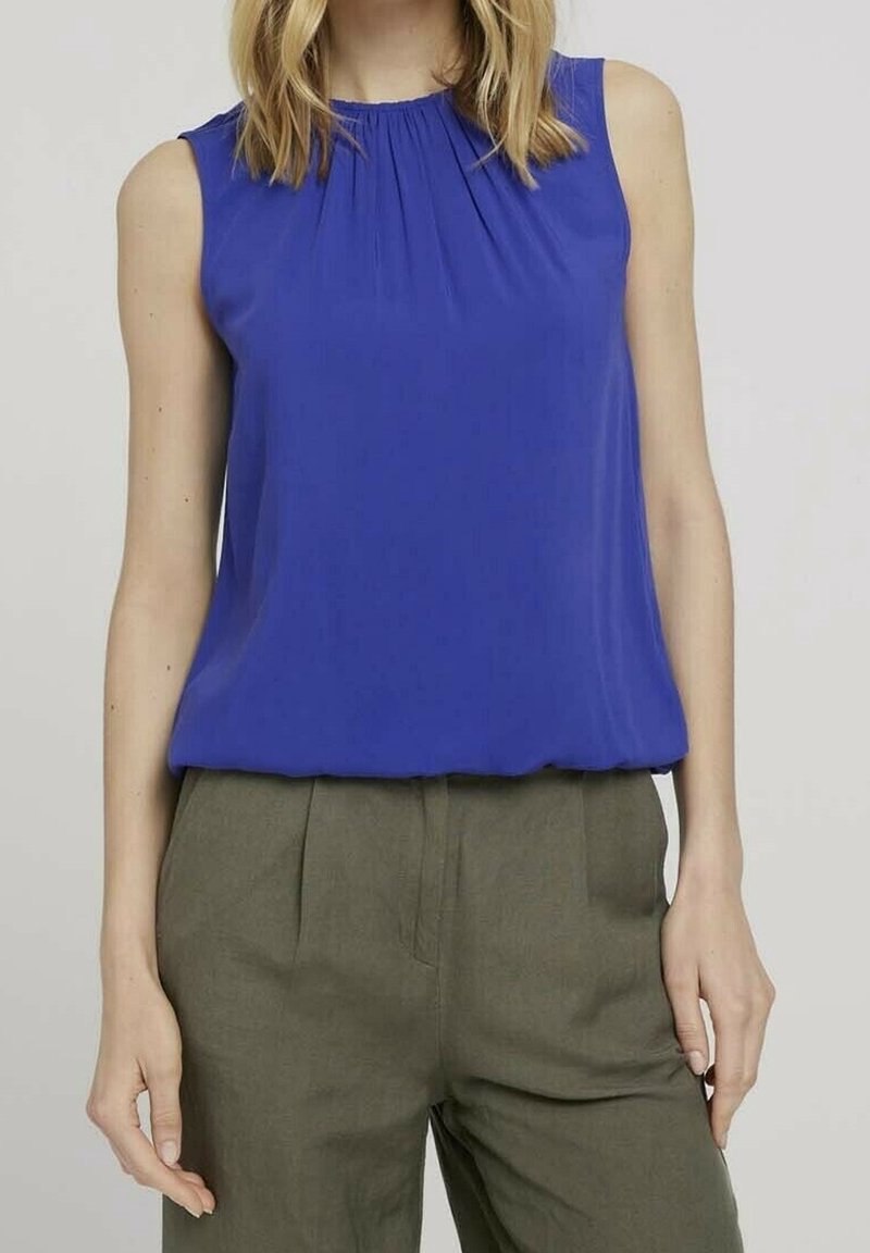 Femme portant un chemisier bleu roi sans manches et un pantalon taille haute vert olive, debout devant un fond uni.