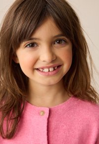 Hellrosa gestrickte Strickjacke mit Rundhalsausschnitt, versehen mit einem einzelnen Knopfverschluss und weicher Textur. Braune Haare umrahmen das Gesicht.