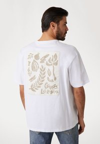 Hvid bomulds t-shirt med et tekstureret beige grafisk design på ryggen, der viser tropiske blade og teksten "Guess Los Angeles".