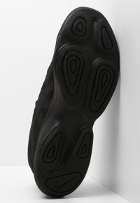 Suela de zapatilla negra con patrones circulares texturizados y el logo de Nike visible en el talón, inclinada contra un fondo de pared blanca en esquina.