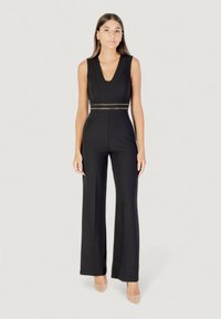 Mono negro con un profundo escote en V, cuerpo ajustado y pantalones de pierna ancha. Cinturón con detalles dorados que resalta. Tejido suave y diseño minimalista.