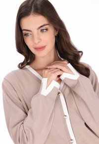 Bézs kötött kardigán fehér peremmel, V-nyakkivágással és arany gombokkal. Hosszú ujjú, kontrasztos mandzsettákkal. Puha tapintású és laza szabású.