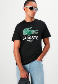 Sort bomulds t-shirt med et grønt krokodillelogo og teksten "LACOSTE PARIS" i hvid. Båret med lyseblå jeans og grå solbriller.