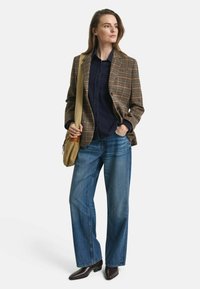 Donna che indossa un blazer a quadri marrone, una camicia blu navy, jeans a gamba larga blu, scarpe marrone scuro e porta una borsa a tracolla color beige, in piedi con una mano in tasca.
