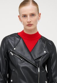 Veste de motard en cuir noir avec matériel en argent, zips diagonaux et texture lisse, superposée à un pull à col roulé côtelé rouge.