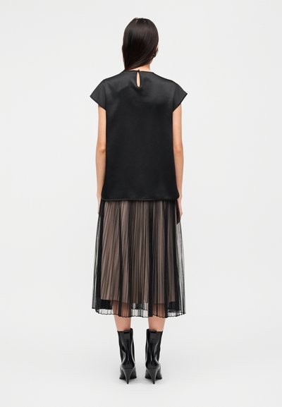 Μαύρο τοπ με κοντά μανίκια και άνοιγμα στο πίσω μέρος, συνδυασμένο με πλισέ φούστα σε μαύρο και γκρι χρώμα. Το μοντέλο φοράει μαύρες μυτερές μπότες.