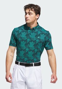 Groene polo met een geometrisch patroon, korte mouwen en een kraag; gecombineerd met witte broek en een zwarte riem.