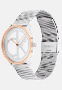 Sidabrinis metalinis laikrodis su apvaliu korpusu, rožinio aukso akcentais, baltu ciferblatu ir tinkline apyranke. Pasižymi minimalistiniu dizainu su ryškiu CK logotipu.