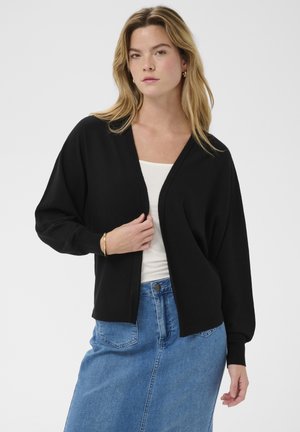 Nő fekete nyitott kardigánban, fehér felsőben, kék farmer szoknyában, arany karkötővel és karika fülbevalókkal, egyszínű háttér előtt pózol.