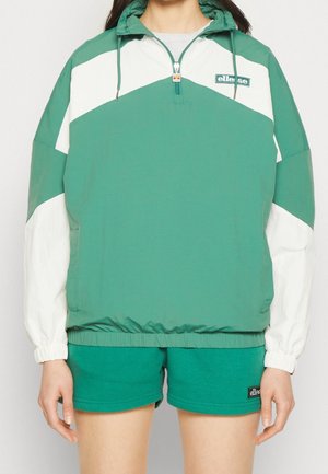 Personne portant une veste mi-zip verte et blanche avec capuche et un short vert assorti sur un fond clair uni.