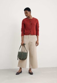 Rdeč kardigan z teksturo in gumbi, bež široke hlače iz kordoroya, zeleni usnjeni crossbody nahrbtnik in temni balerinki. Enostaven in čist dizajn.