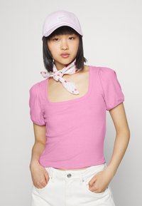 Top rosa com textura, de mangas curtas e decote quadrado, combinado com um lenço branco com detalhes em vermelho e um boné lilás claro.