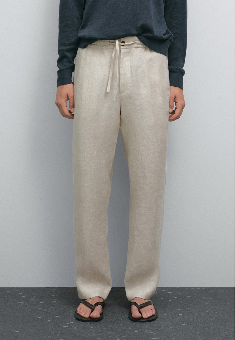 Personne portant un pantalon en lin beige avec cordon, des tongs noires et un haut à manches longues foncé, debout sur un sol foncé tacheté.