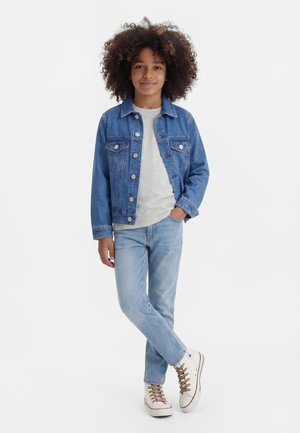 Kind mit lockigem Haar, das eine blaue Jeansjacke, ein hellgraues Hemd, hellblaue Jeans und weiße Turnschuhe mit bunten Schnürsenkeln trägt und im Schneidersitz dasteht.