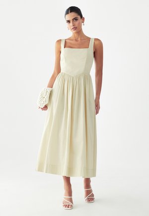 Femme portant une robe midi crème sans manches avec un col carré, tenant une pochette tissée et chaussée de talons blancs à brides, se tenant devant un fond uni.