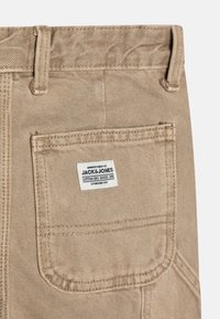 Pantaloni in cotone beige con una tasca posteriore dotata di etichetta, un tessuto testurizzato con cuciture visibili e una finitura spazzolata.