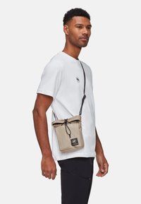 Sac bandoulière beige avec fermeture à cordon, bandoulière réglable et patch logo. Porté sur un t-shirt blanc, avec une forme rectangulaire élégante.