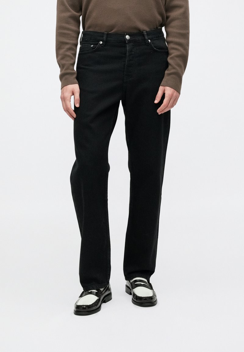 Vaqueros de mezclilla negra con un diseño de pierna recta, cinco bolsillos, cierre de botón y sutiles detalles de costura. Llevados con mocasines en blanco y negro.