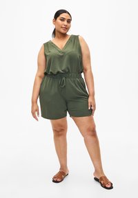 Salopette senza maniche verde oliva con scollo a V, dettagli arricciati, vita elastica e pantaloni corti in tessuto abbinato. Sandali aperti marroni.