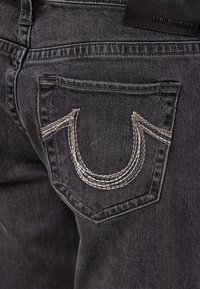 Jeans de mezclilla negra con un bolsillo trasero distintivo que presenta un diseño en forma de U bordado en gris y costuras en contraste. La etiqueta dice "True Religion."