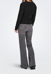 Femme debout, légèrement de profil, portant un haut noir ajusté à manches longues et un jean gris droit avec poches arrière et des chaussures noires.