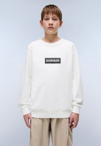 Sudadera blanca de manga larga y cuello redondo, con un parche de logo rectangular negro centrado en el pecho. Pantalones beige claros visibles.