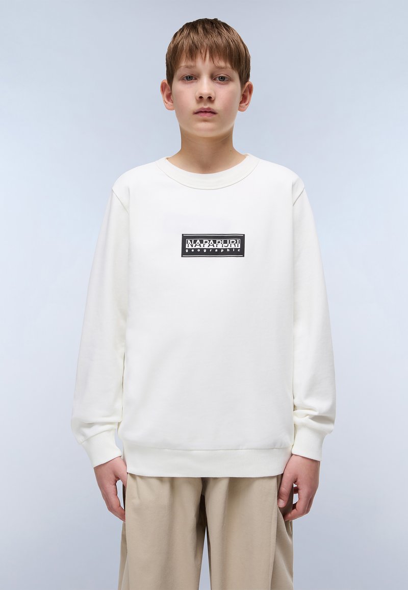 Sudadera blanca de manga larga y cuello redondo, con un parche de logo rectangular negro centrado en el pecho. Pantalones beige claros visibles.