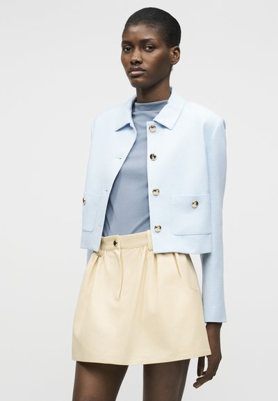 Γυναίκα ντυμένη με ανοιχτόγαλανη κοντή ζακέτα με χρυσά κουμπιά πάνω από μπλε τοπ και μπεζ φούστα ψηλόμεση, που στέκεται σε απλό φόντο.