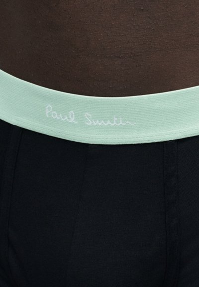 Gros plan sur une taille à peau foncée portant des sous-vêtements noirs avec une ceinture vert clair et un logo blanc "Paul Smith".