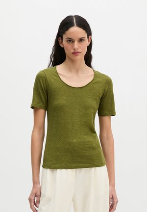 Femme aux longs cheveux foncés portant un haut vert olive à manches courtes et col rond et un pantalon blanc ample devant un fond uni.