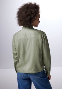 Una camicia verde oliva a maniche lunghe con una texture liscia, polsini con bottoni e un dettaglio sul retro. Abbinata a jeans di denim blu.