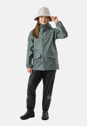 Giovane persona che indossa un cappello da pescatore grigio chiaro, giacca antipioggia verde con tasche a scatto, pantaloni impermeabili neri e stivali neri, in piedi su sfondo bianco.