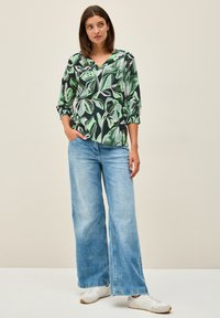Haut à imprimé floral en vert, noir et blanc, avec un décolleté en V et des manches 3/4. Assorti à un jean large bleu clair et des baskets blanches.