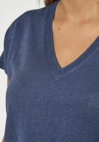 Mornarsko plava V-majica od teksturiranog materijala s laganim sjajem i kratkim rukavima, prikazuje dio dekoltea.